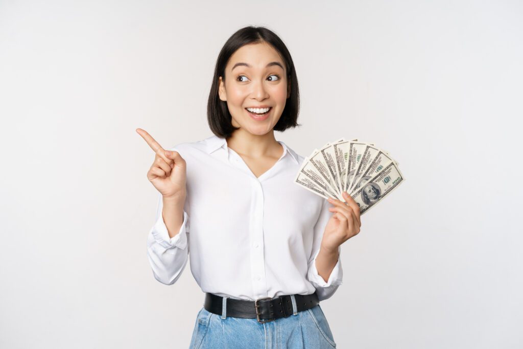 smiling young modern asian woman pointing banner advertisement holding cash money dollars standing white background